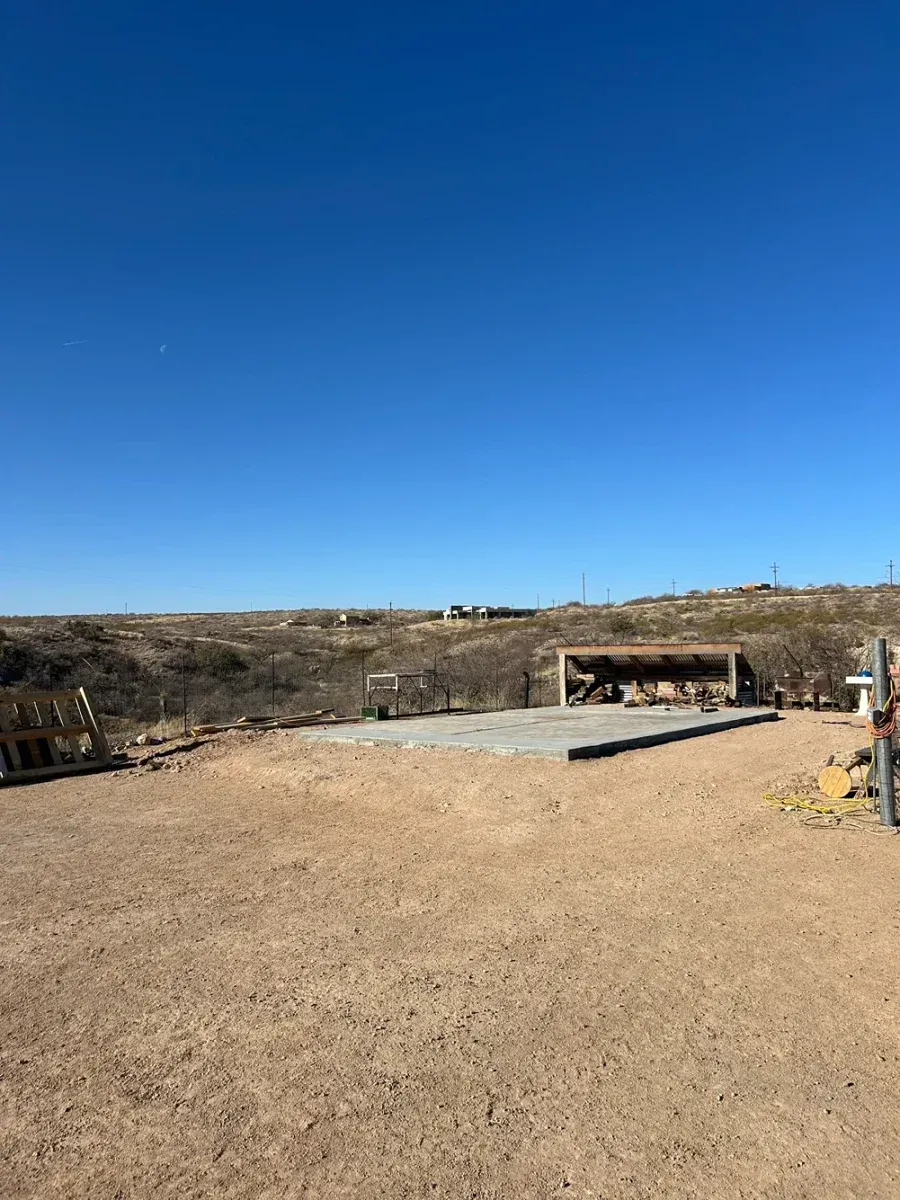 Wide dirt lot with a concrete pad and hillside under a clear blue sky