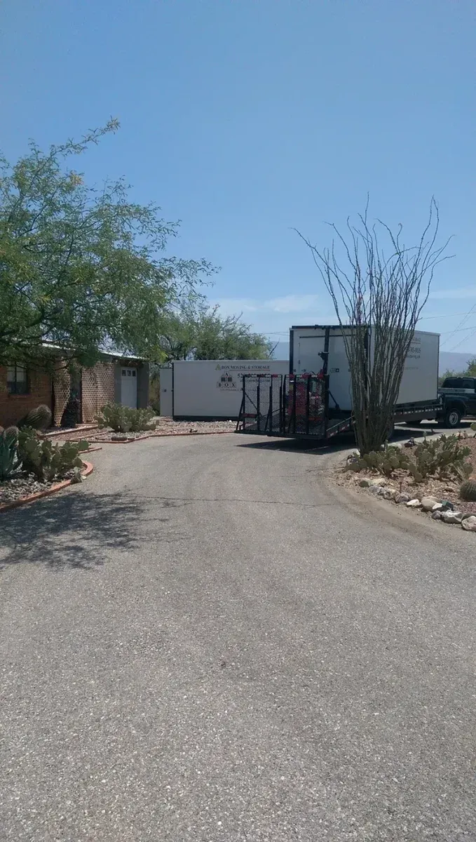 Wide gravel driveway leading to a white building and trailer, with trees and desert landscaping on both sides