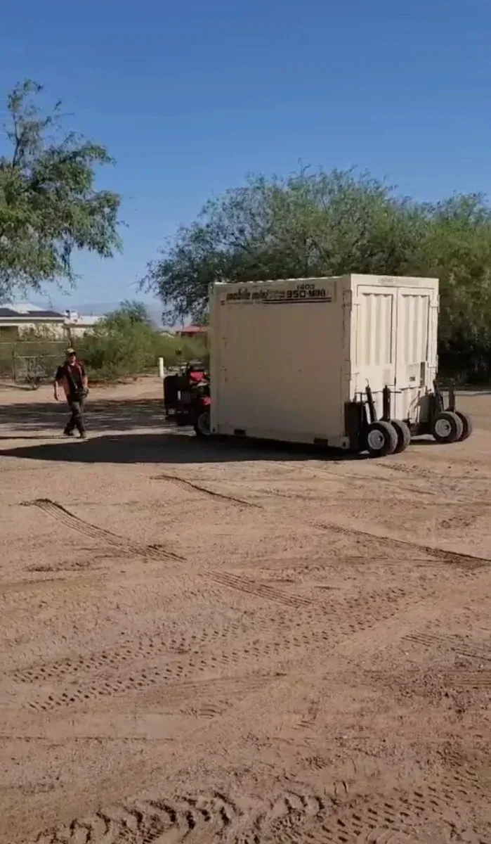 Beige box trailer parked on a dirt road beside a person and motorcycle, with sparse trees and blue sky.