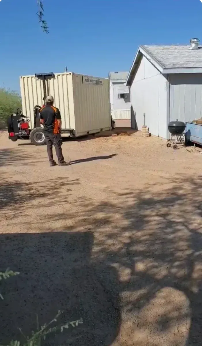 Person standing in a dusty yard near a beige container and a white building under a blue sky