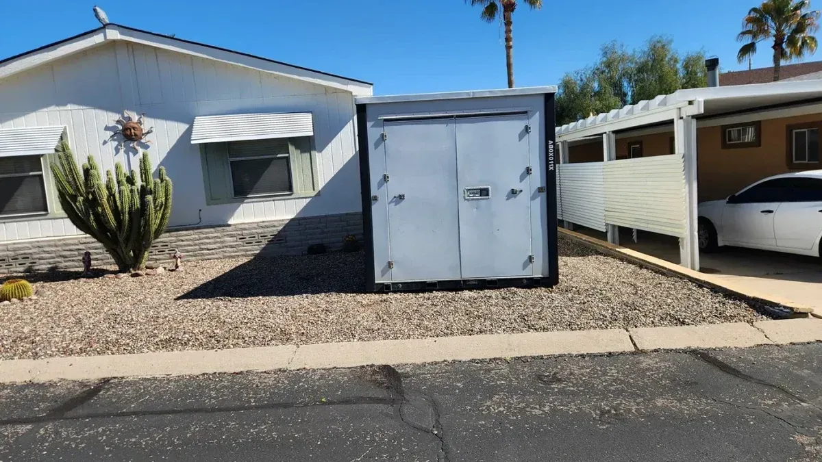 White garage-style storage container on a gravel driveway in front of a suburban house under a blue sky.