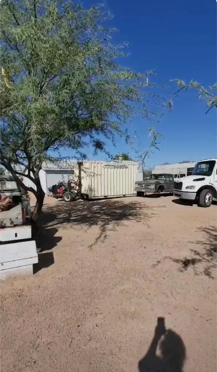 Desert campsite with trees, a white storage container, parked trucks, and a clear blue sky