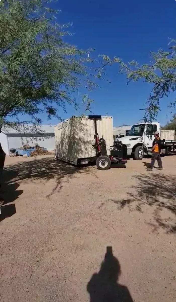 Workers beside a white truck and equipment trailer in a dusty lot under a clear blue sky.