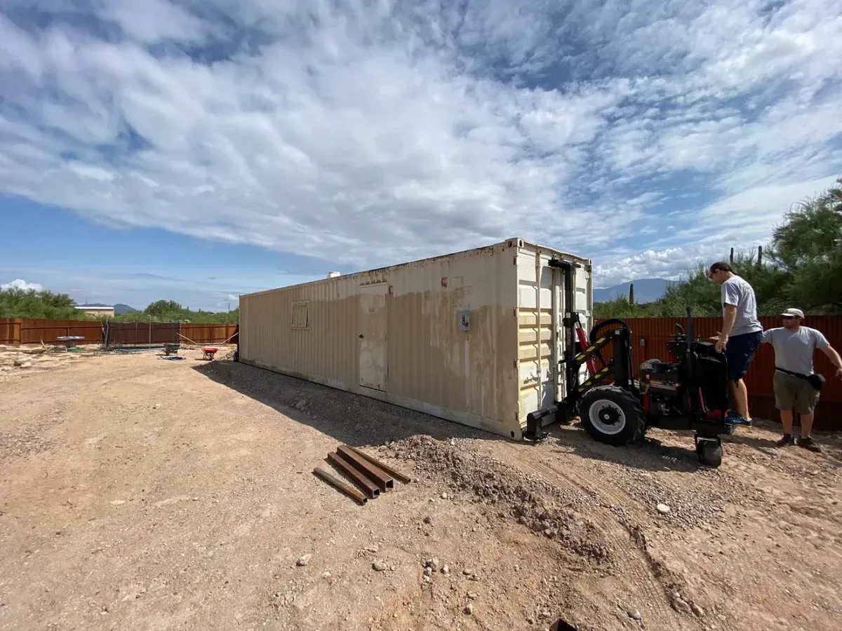 Shipping container and small tractor on a dirt lot under a cloudy sky, with two people nearby
