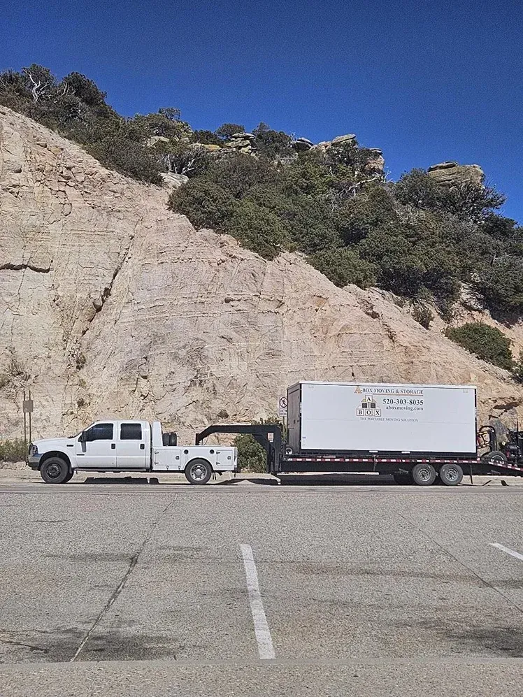 White pickup truck towing a white box trailer parked by a rocky hillside under a clear blue sky