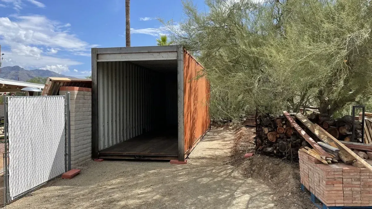 Open shipping container in a dusty yard beside fencing, trees, and stacked lumber.