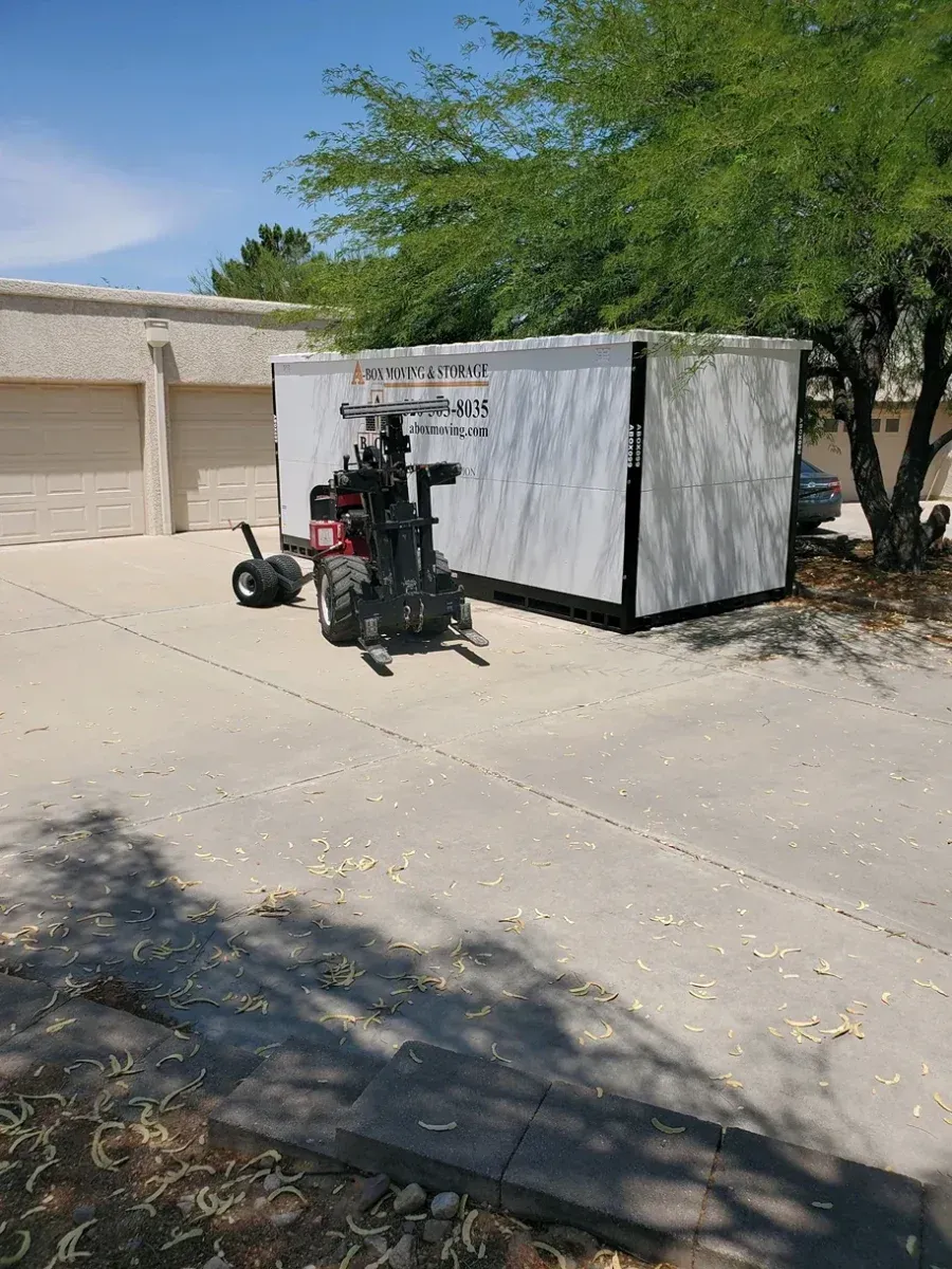 Motorized wheelchair parked beside a white storage container in a sunny driveway under a tree.
