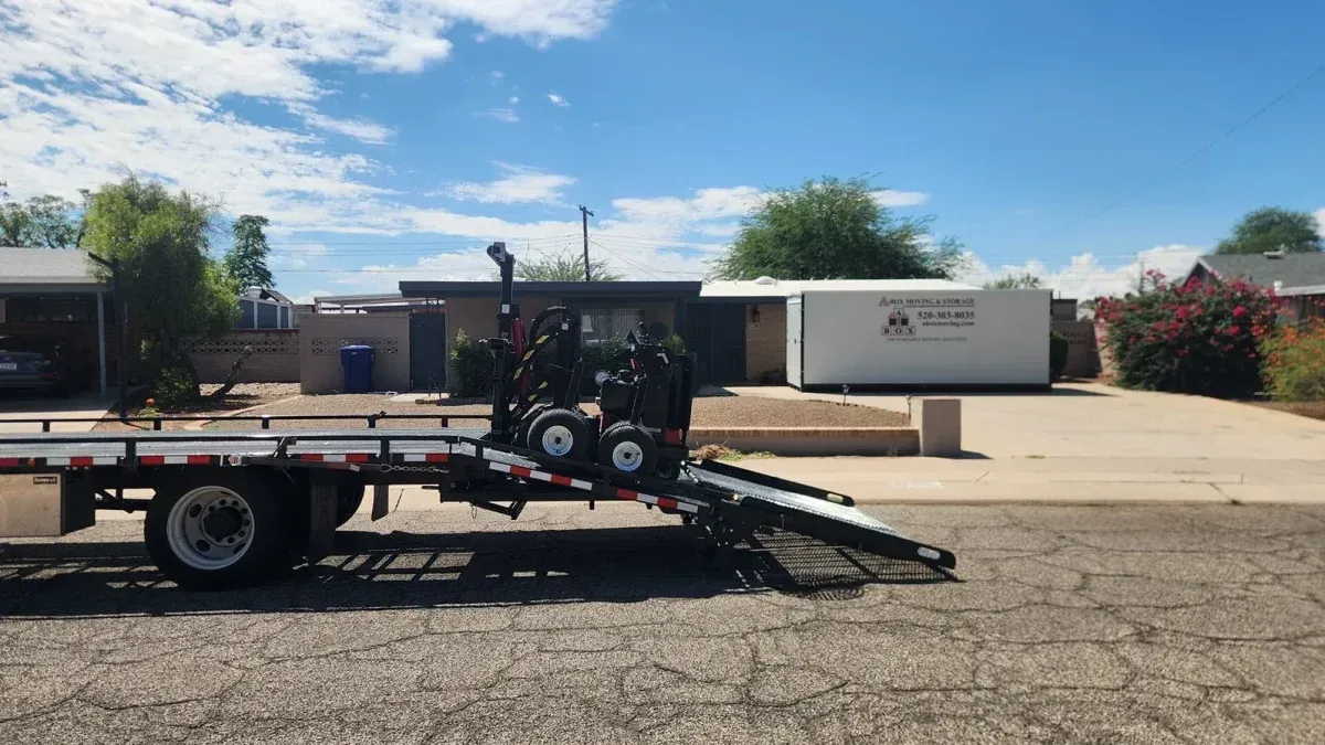 Trailer with lawn equipment parked in a sunny lot beside a white truck and buildings