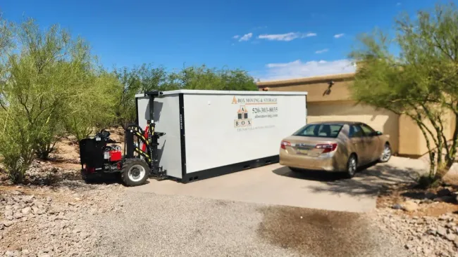 Small white trailer and forklift beside a tan car in a desert yard near a house, with trees and blue sky.