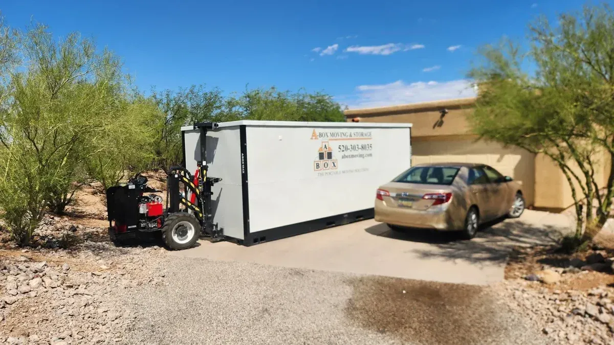 Small trailer and tan car parked on a gravel driveway beside a beige house in a desert yard.