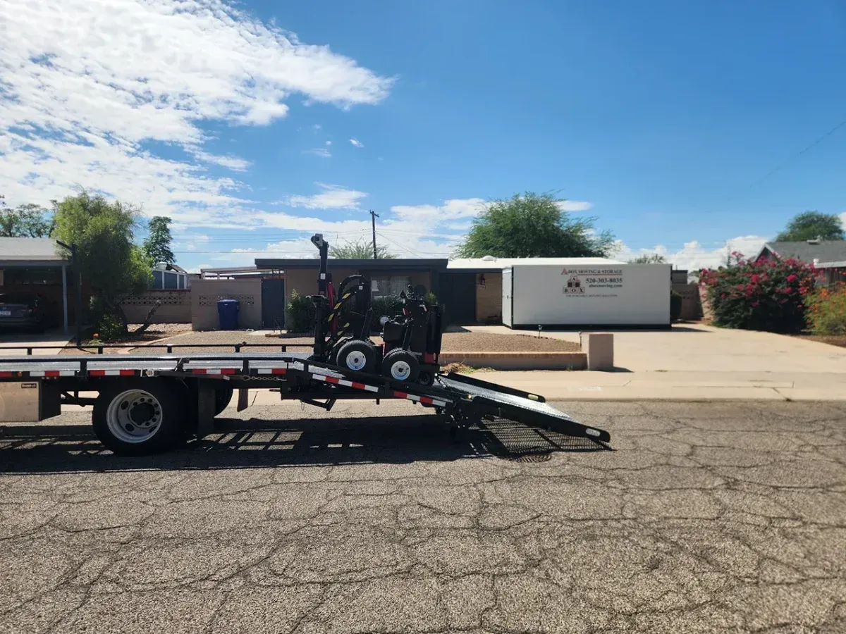 Flatbed trailer with a ramp parked in a cracked lot under a blue sky, with a white container in the background.