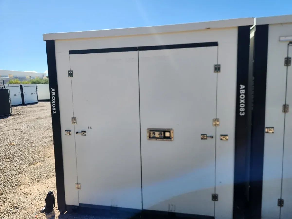 White outdoor utility cabinet with double doors and black trim in a gravel lot, beside another cabinet.