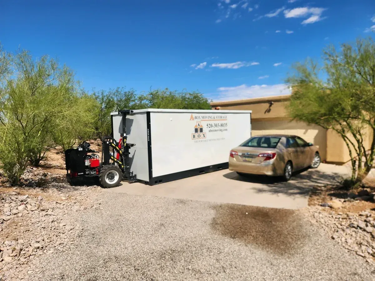 White utility trailer and small tractor beside a tan car on a gravel driveway near a desert house.