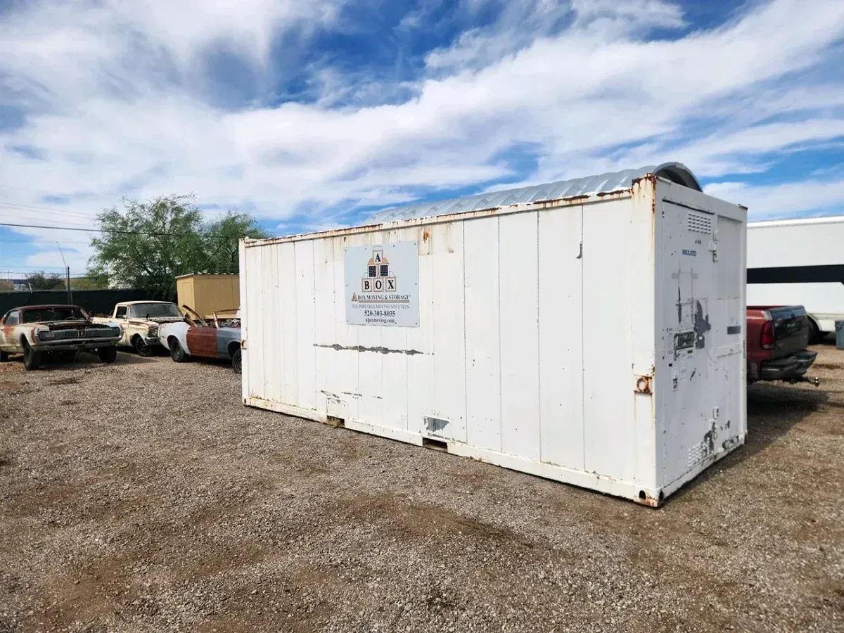 White shipping container in a gravel lot with parked vehicles under a cloudy sky