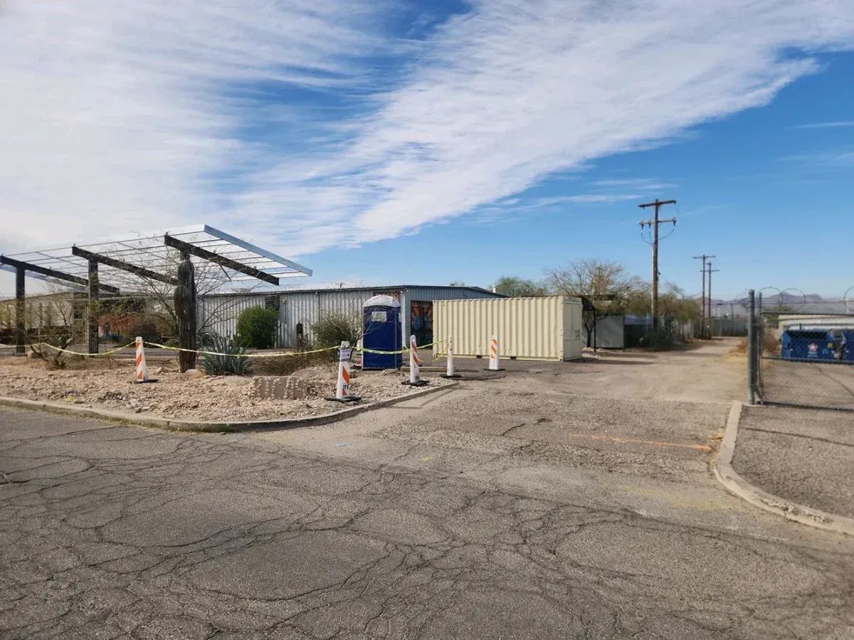 Desert roadside with a small building, blue portable restroom, and metal canopy under a cloudy sky