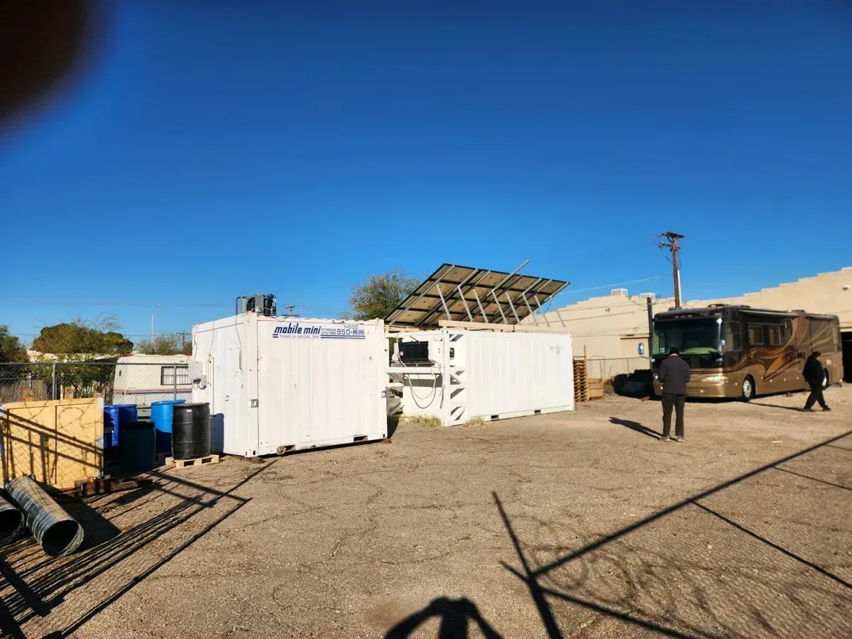 Desert site with white modular buildings, solar panels, and two people walking on a dirt lot under a blue sky