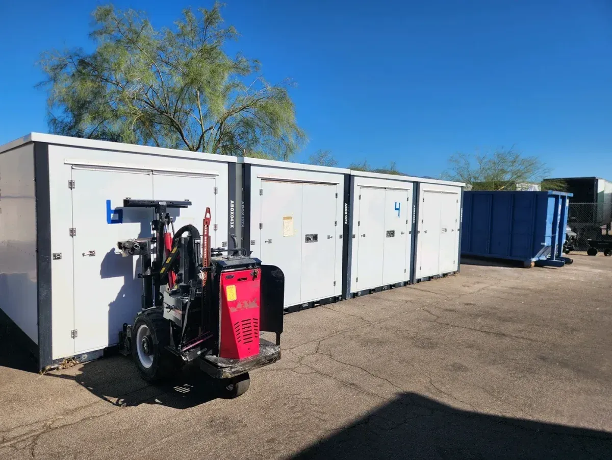 Trailer-mounted pressure washer parked beside white storage containers under a clear blue sky