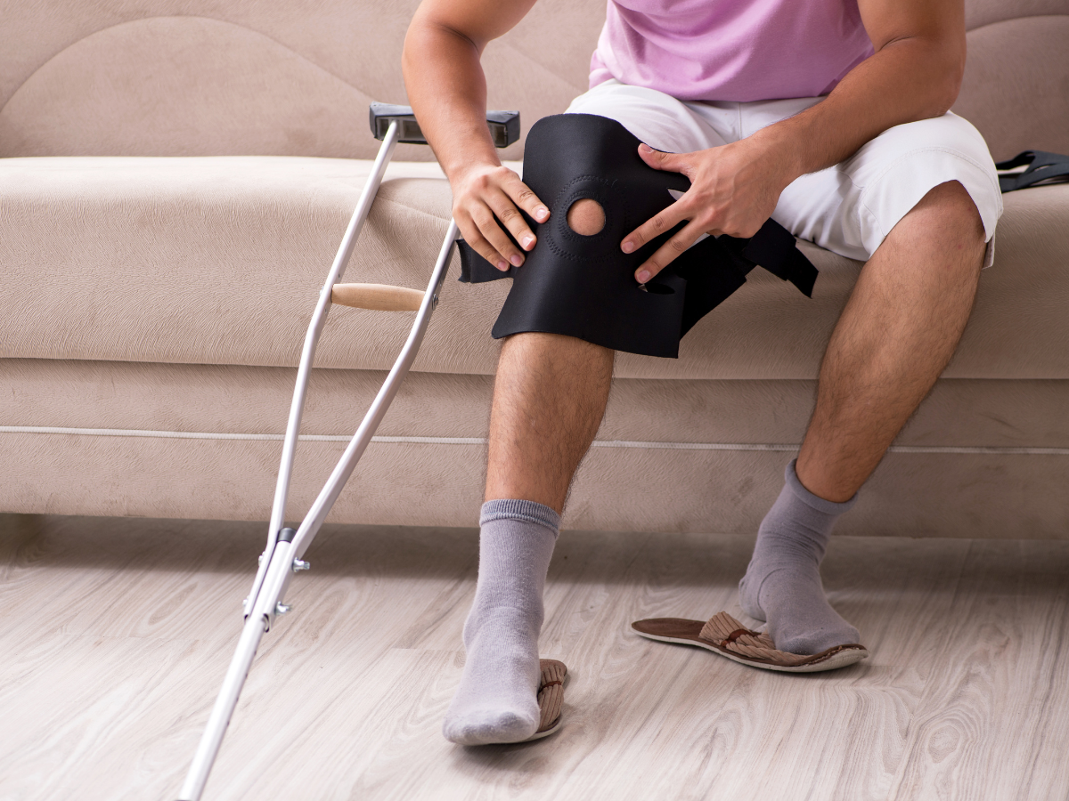 Person sitting on a couch, holding a black knee brace while using crutches on one leg