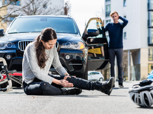Woman sitting on road holding her leg, with a man by an open car door and scattered bicycles nearby