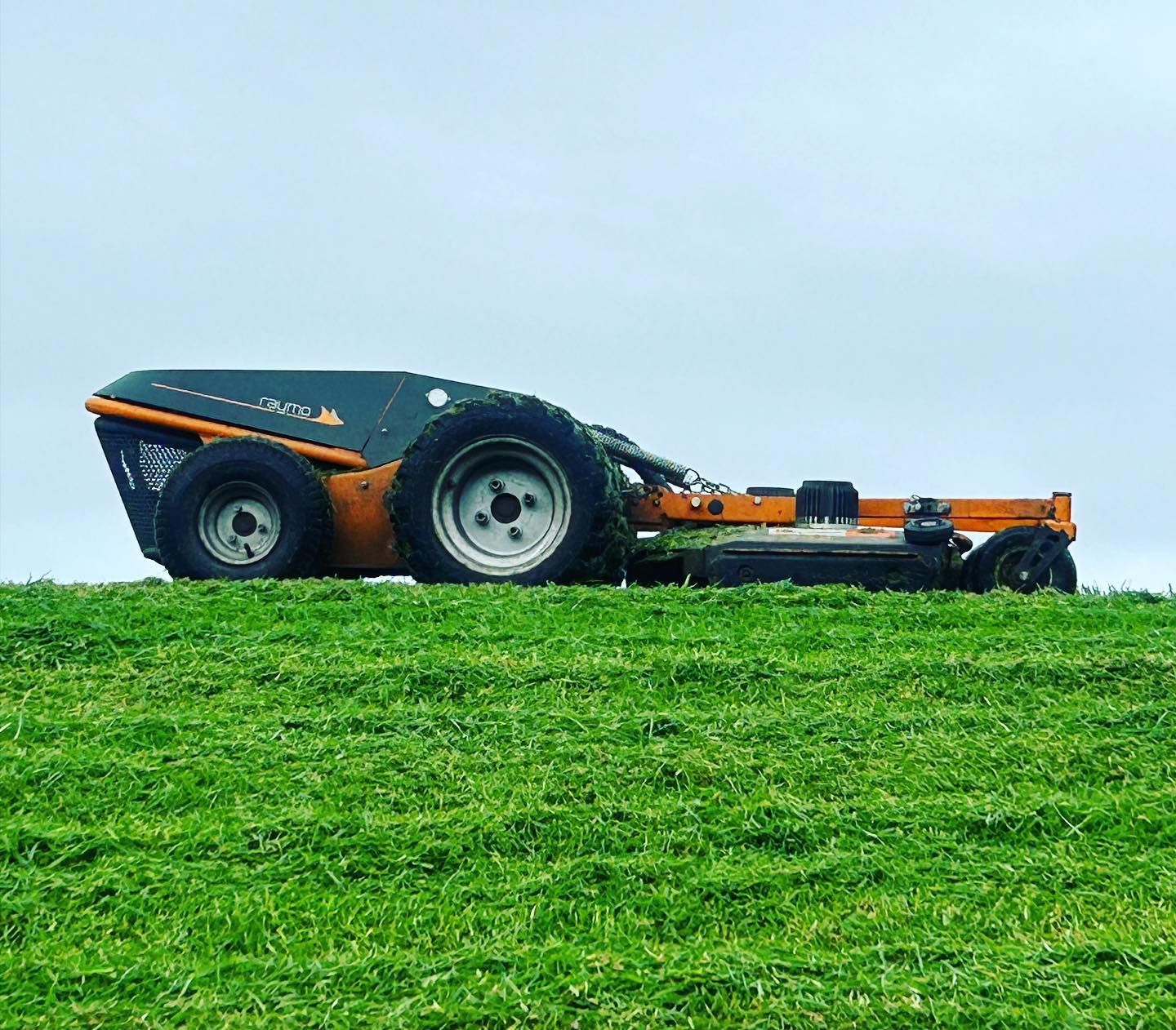 Robotic lawnmower on a green grassy hill, cutting the lawn. Gray and orange machine, cloudy sky.