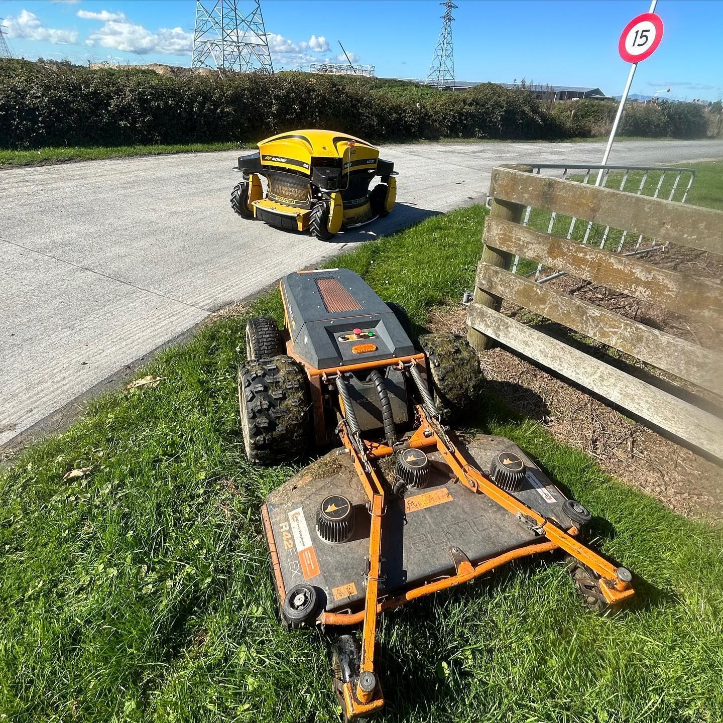 Two robotic mowers: one yellow on a road, one orange/gray mowing grass near a fence.