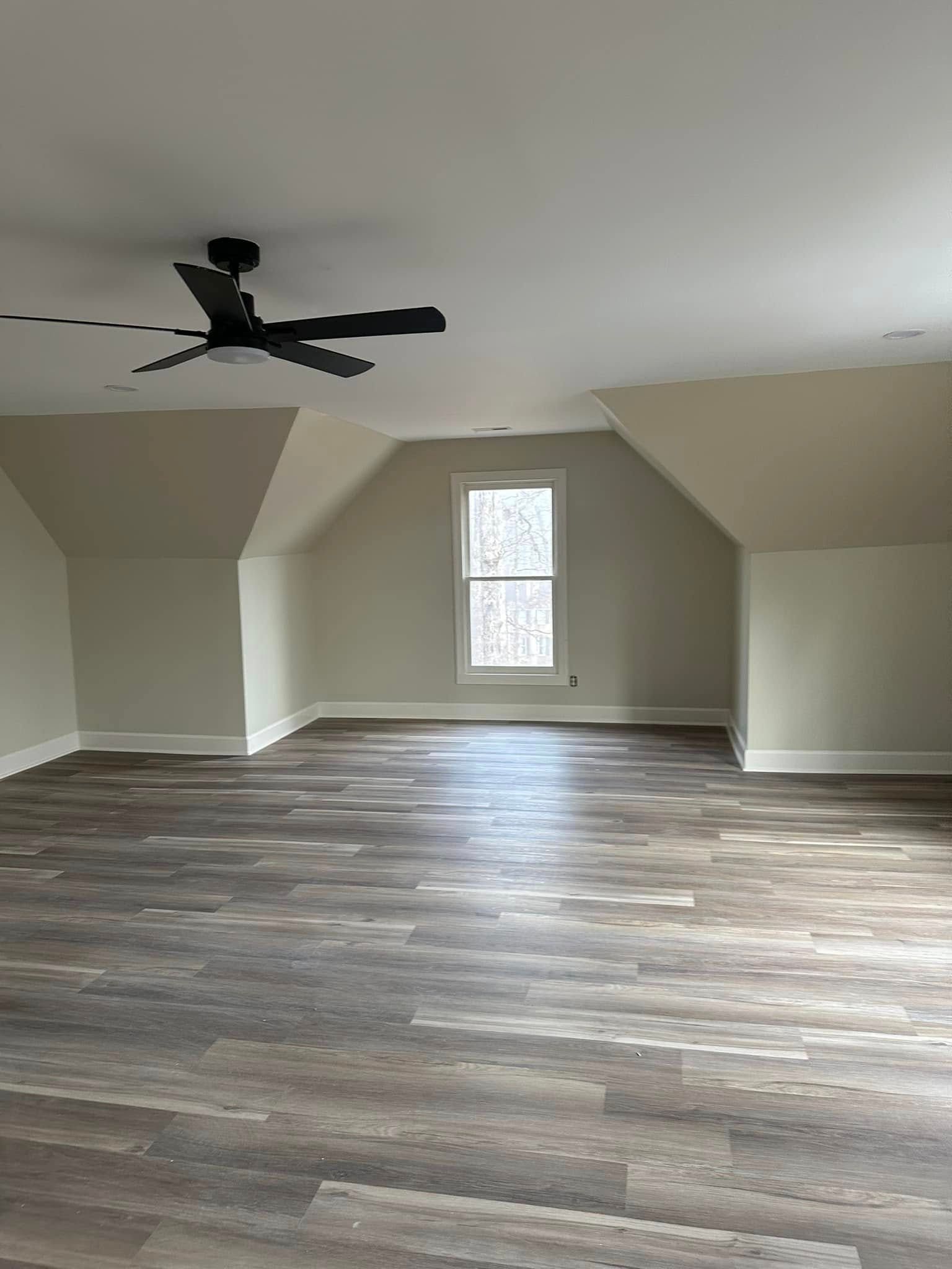 Spacious room with wood-look flooring, sloped ceiling, window, and ceiling fan. Beige walls and white trim.