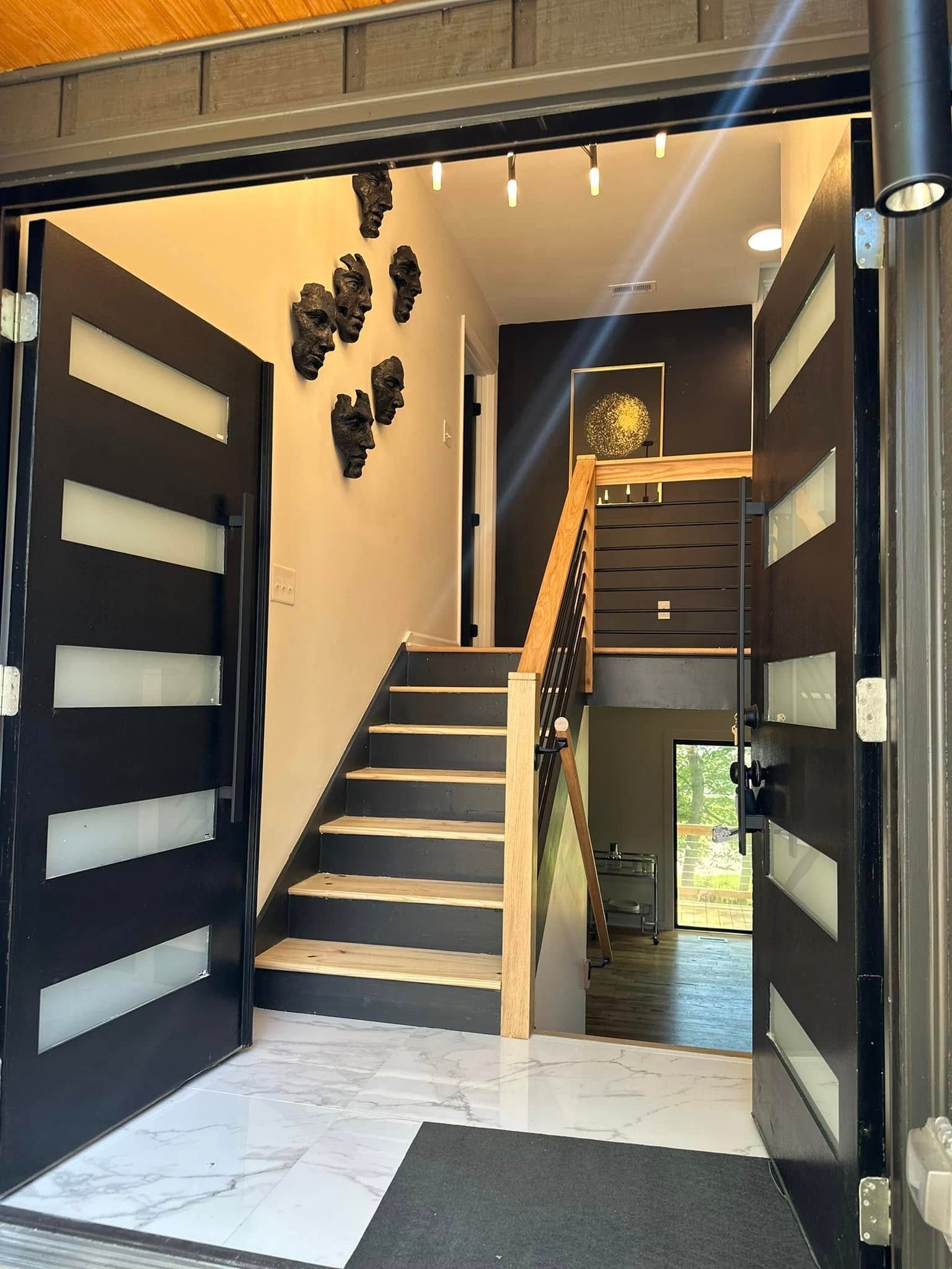Open black front doors reveal a modern entryway with a staircase, artwork, and light fixtures.