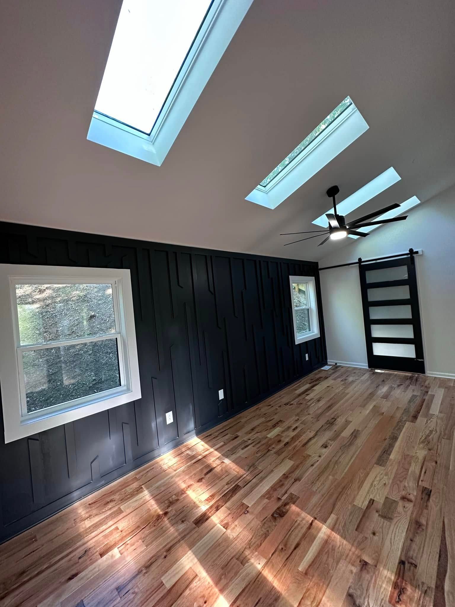 Room with hardwood floors, dark paneled wall, windows, skylights, and a black ceiling fan.