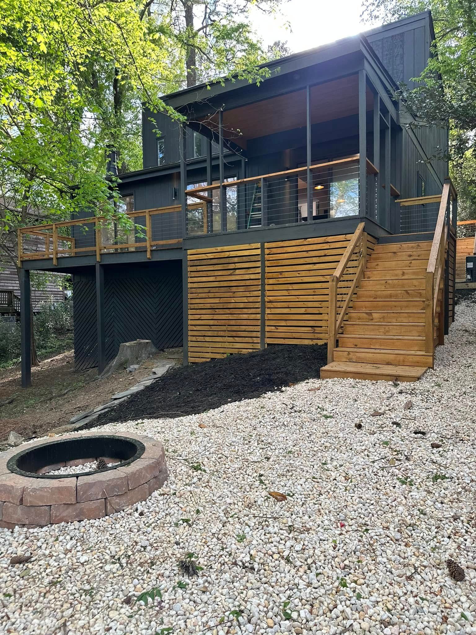 Modern cabin with a deck, stairs, and a fire pit in a wooded area. Black and brown tones.