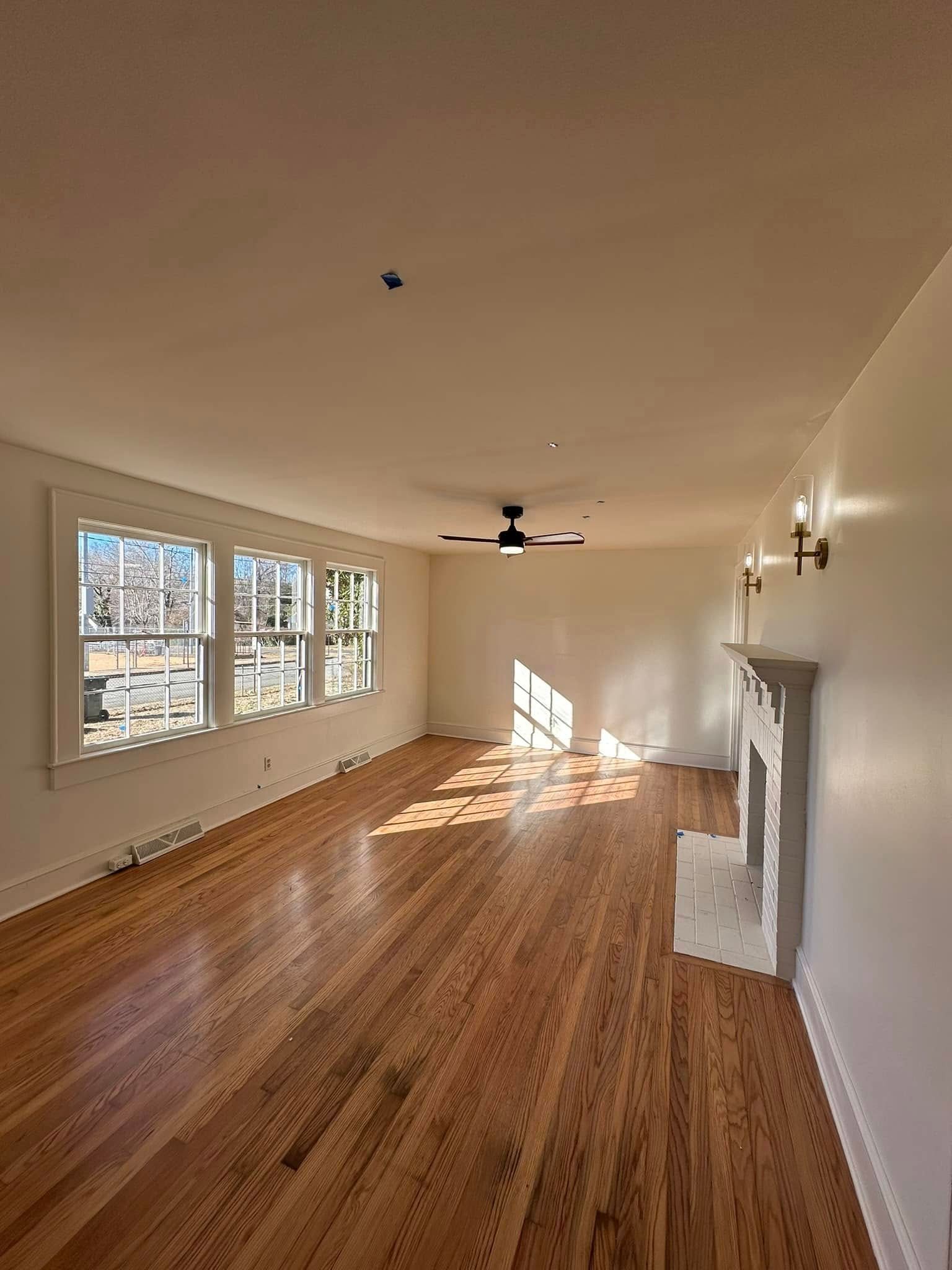 Empty living room with wood floors, windows, fireplace, and ceiling fan.