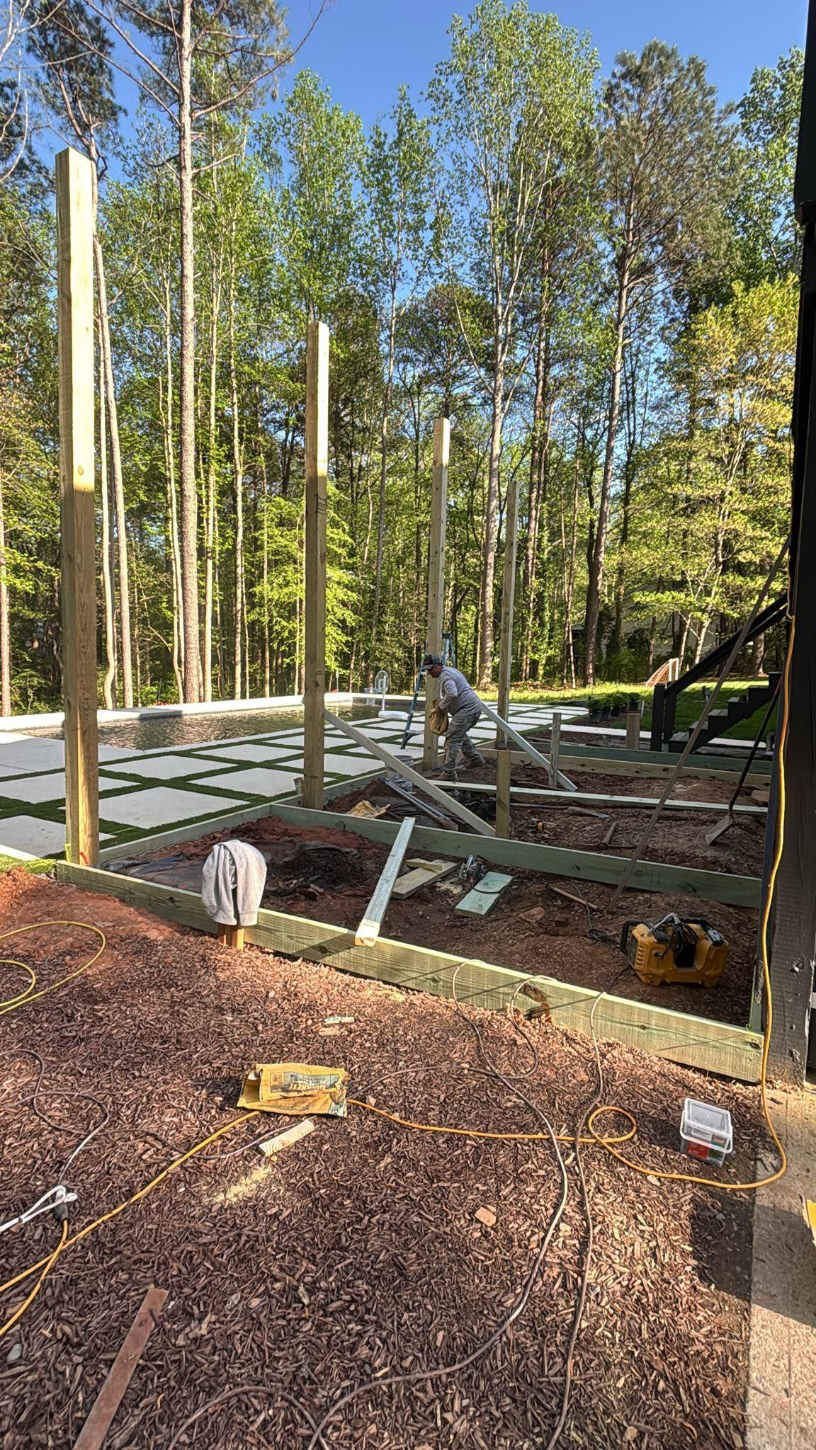 Construction site with wooden posts and boards. A person works amid the building materials in a wooded setting.