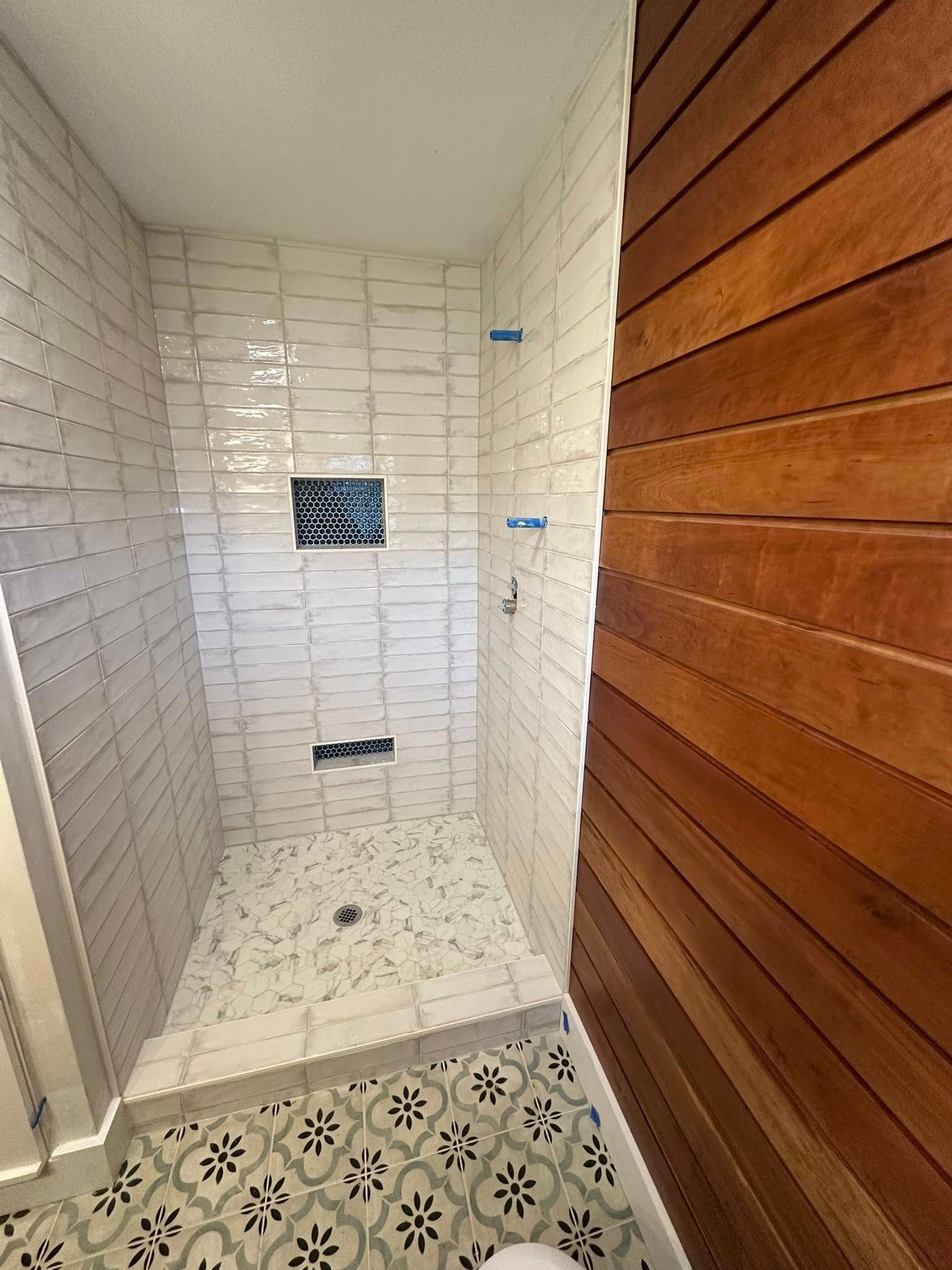Shower stall with white tile walls and patterned floor, wood paneling on the right.