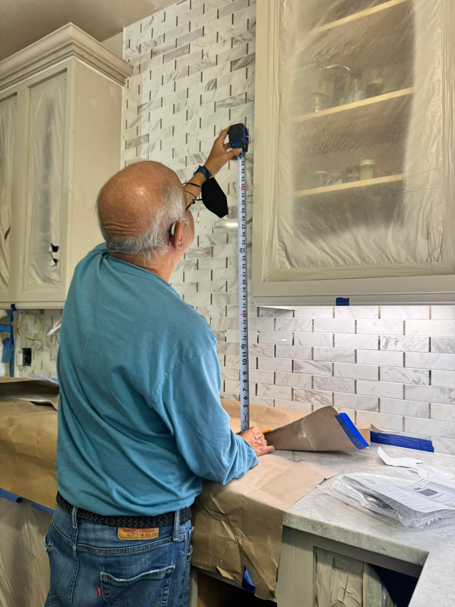 Man measures a backsplash in a kitchen. He wears a blue shirt and jeans, near covered cabinets.