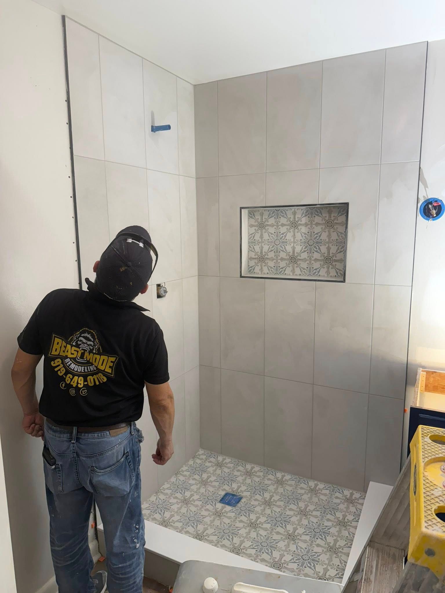 Man tiling a shower stall; gray tile, inset shelf, patterned floor tile.