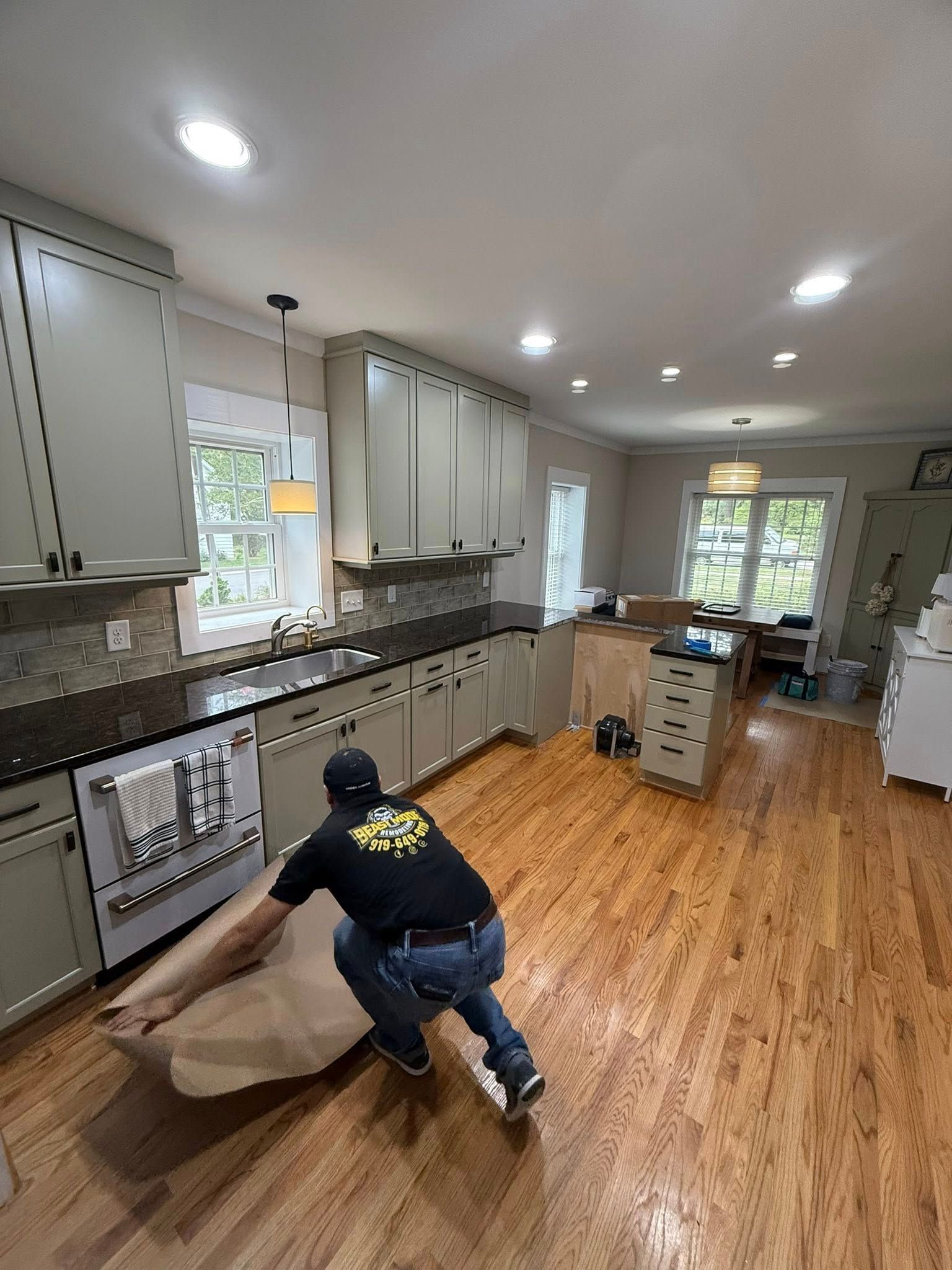Man in a kitchen carrying a wrapped appliance, light gray cabinets, hardwood floors, recessed lighting.