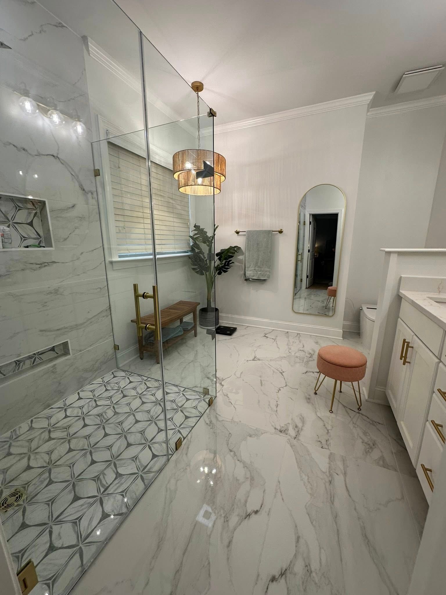 Luxurious white marble bathroom with glass shower, gold accents, and a pink stool.