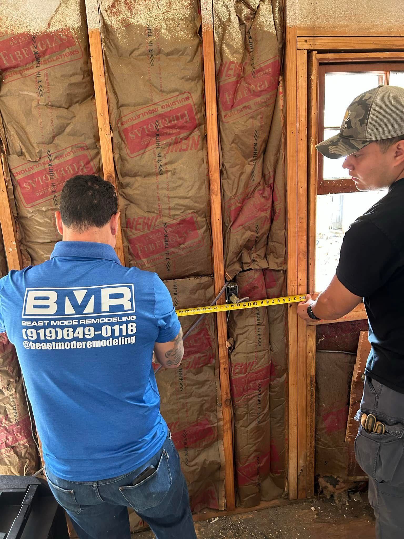 Two people measuring a wall with exposed insulation. One in a blue shirt is holding the tape measure.