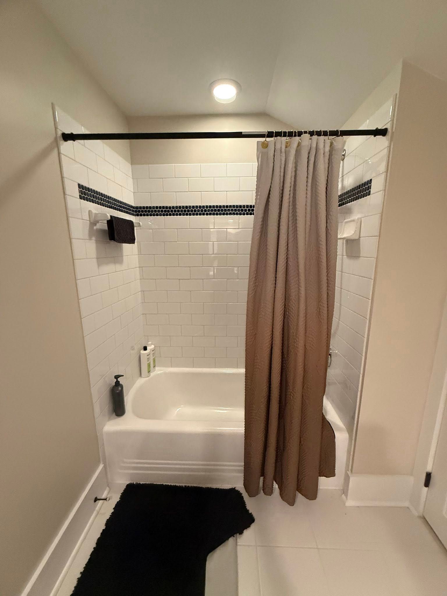 Bathroom with white tiled walls, black trim, a bathtub, and a brown shower curtain. A black bathmat is on the floor.
