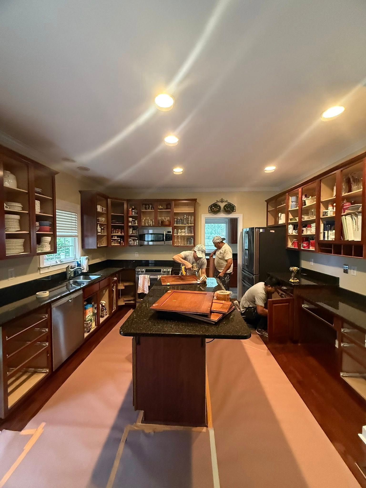 Kitchen undergoing renovation. Cabinets, island, and appliances. Workers visible. Pink floor covering.