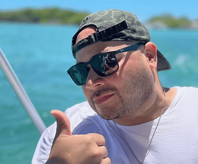 Man wearing sunglasses and camouflage cap gives a thumbs-up on a boat; ocean background.