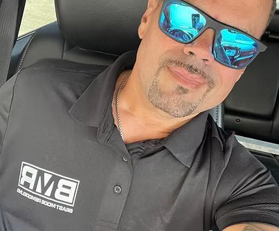 Man wearing sunglasses and a black polo shirt, sitting in a car, smiling.