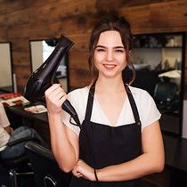 Portrait of smiling hairdresser in beauty salon. Beautiful woman in black apron looking at camera and holding professional hair dryer. Beauty and people concept