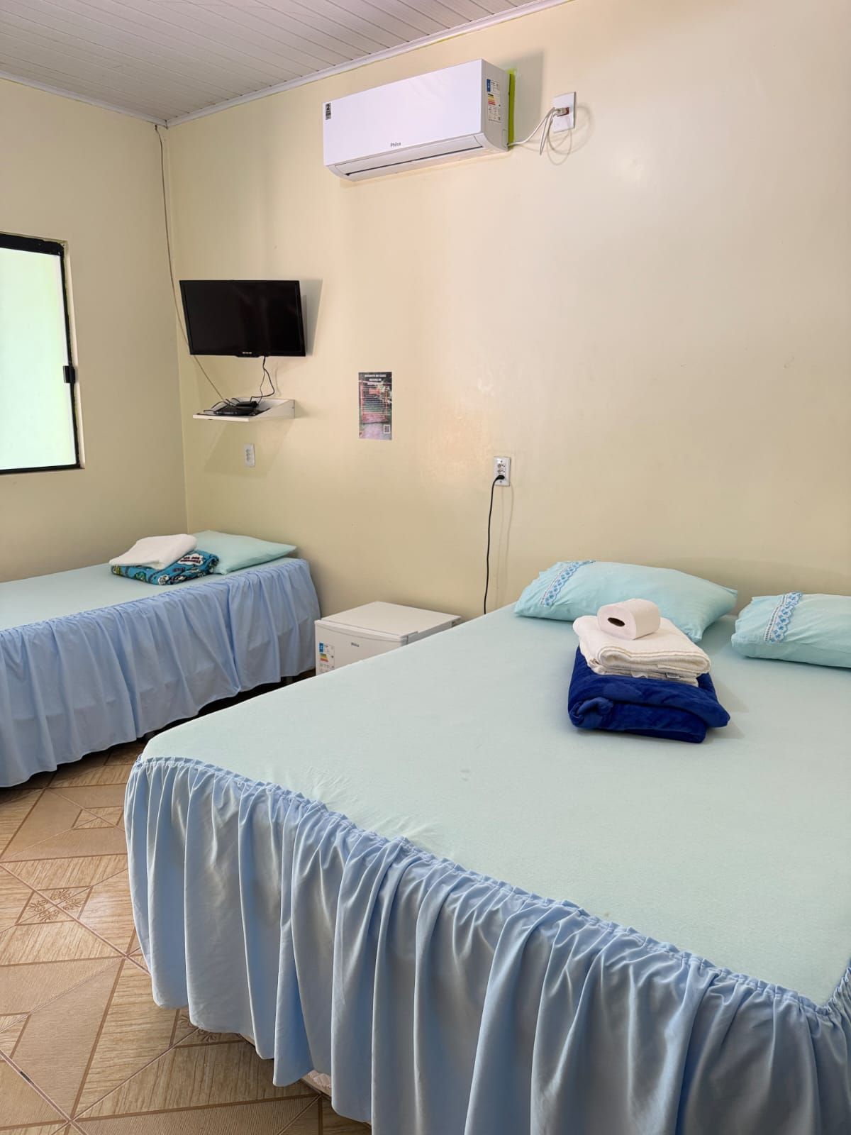 Um quarto de hotel com duas camas azul-claras, uma TV fixada na parede e um ar-condicionado contra uma parede amarelo-clara.