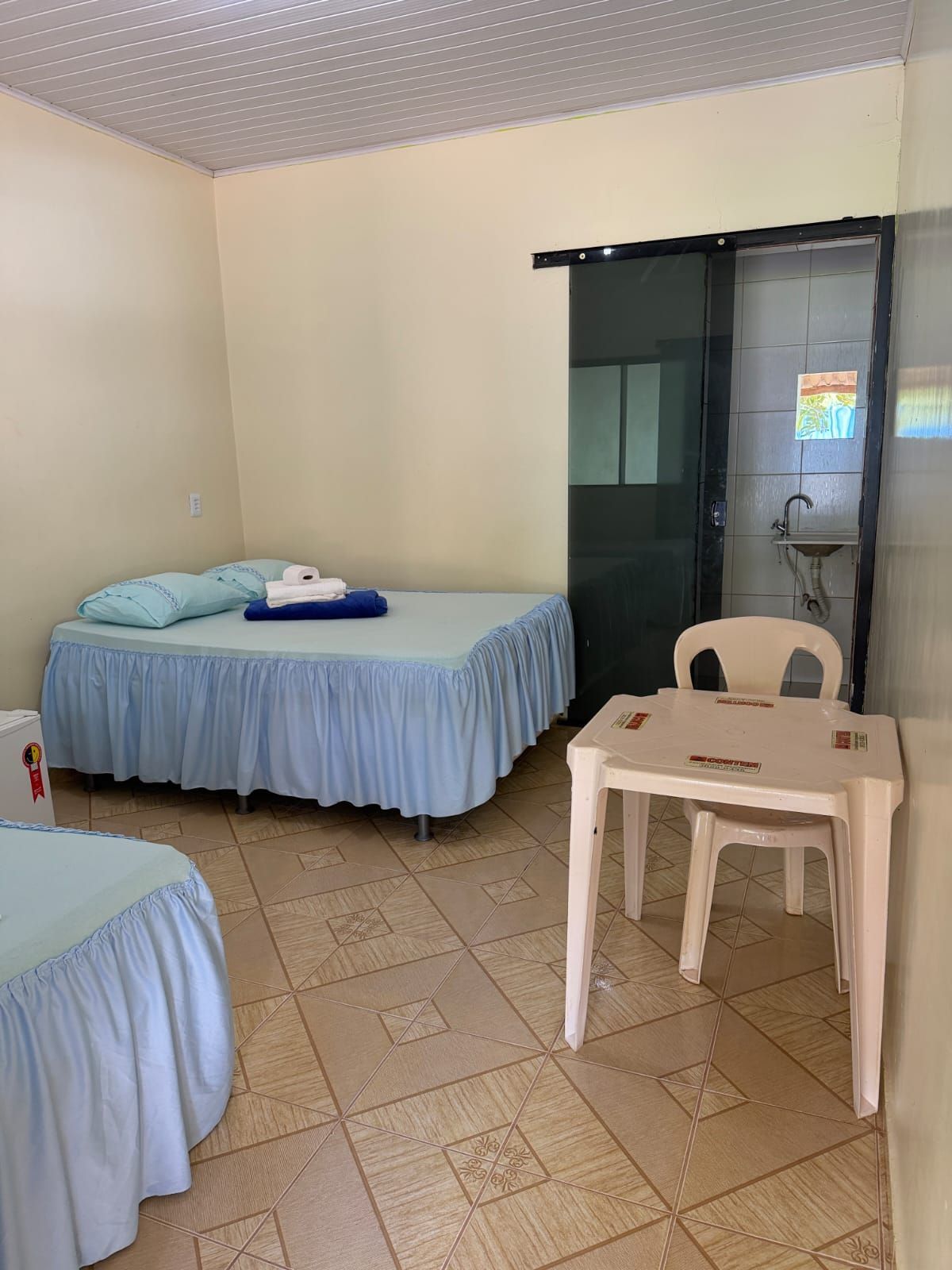 Um quarto de hotel com duas camas com colchas franzidas azul-claras, uma pequena mesa de plástico