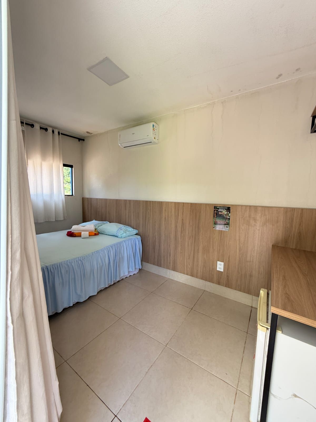 Um quarto com uma cama com saia azul franzida, paredes revestidas de madeira, piso de azulejo e ar-condicionado de parede.