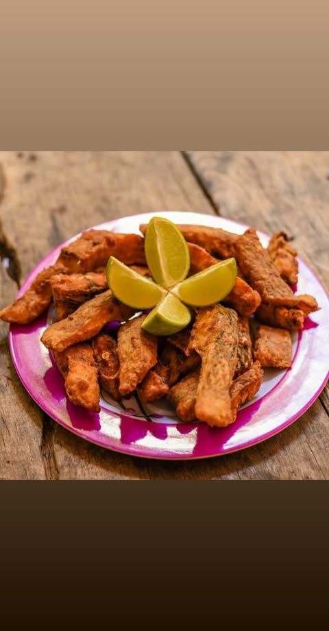 Tiras de peixe frito servidas em um prato rosa com fatias de limão, sobre uma mesa rústica de madeira.