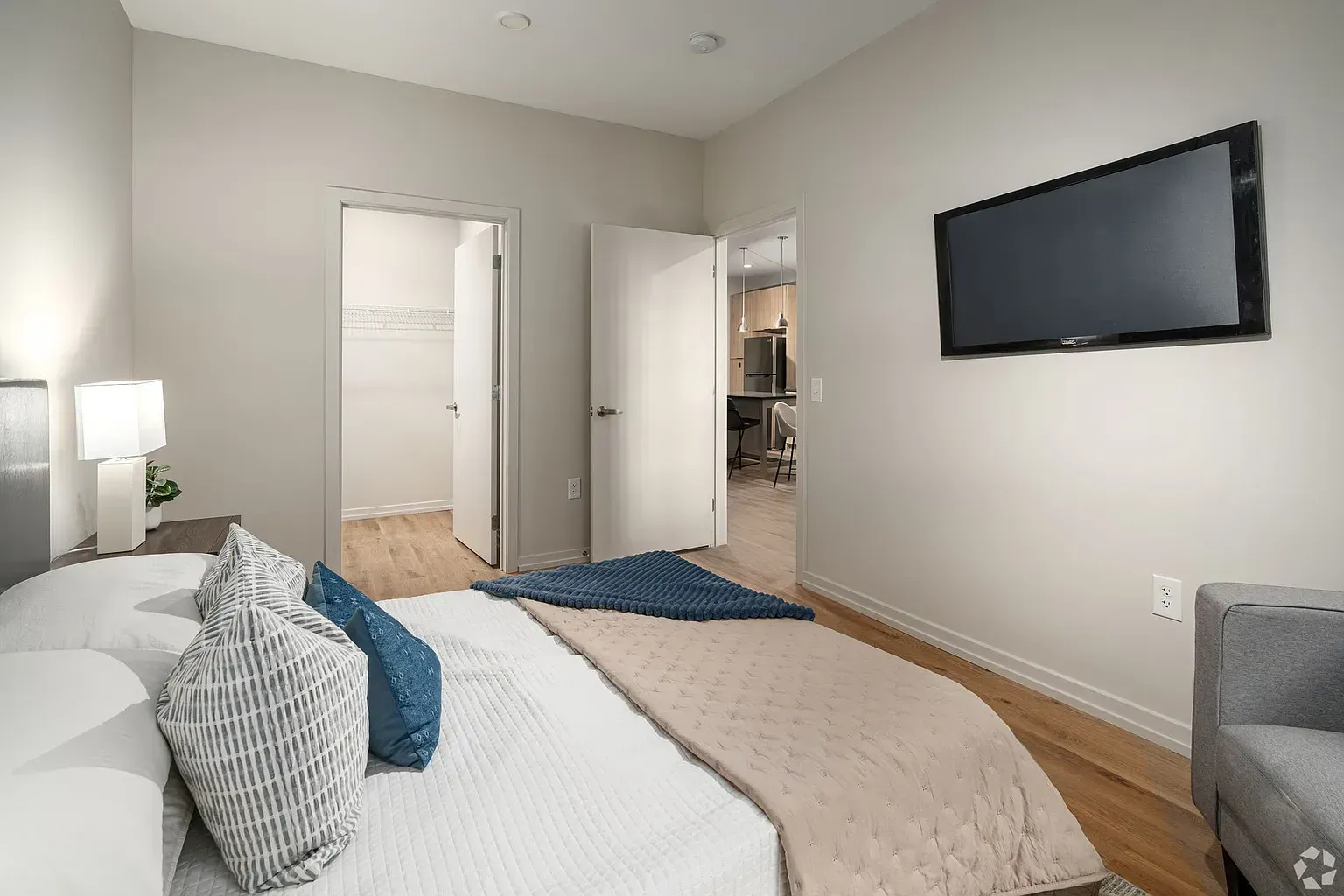 Photo of a bedroom, showing a bed, a tv on the wall, a chair and a nightstand with a lamp on it