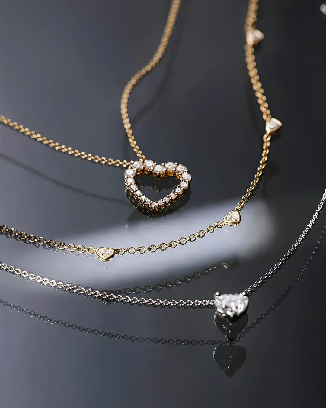 Tre collane a forma di cuore con diamanti su una superficie nera riflettente, catene in oro e argento.