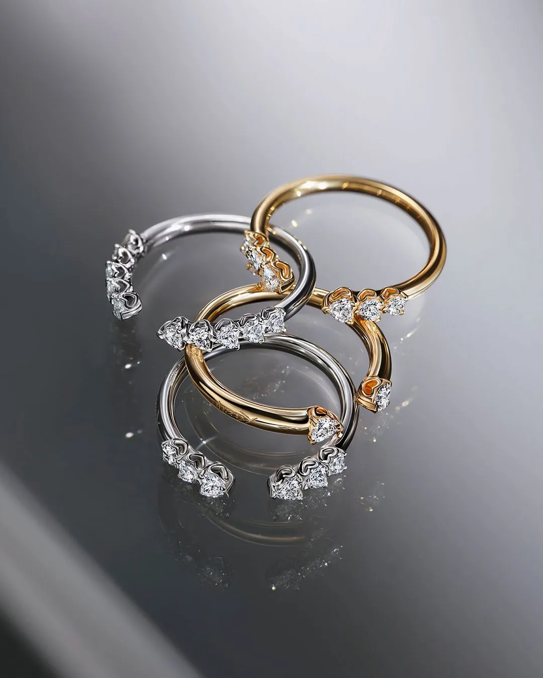 Tre anelli aperti, in oro e argento, con piccoli diamanti incastonati alle estremità, su una superficie grigia riflettente.