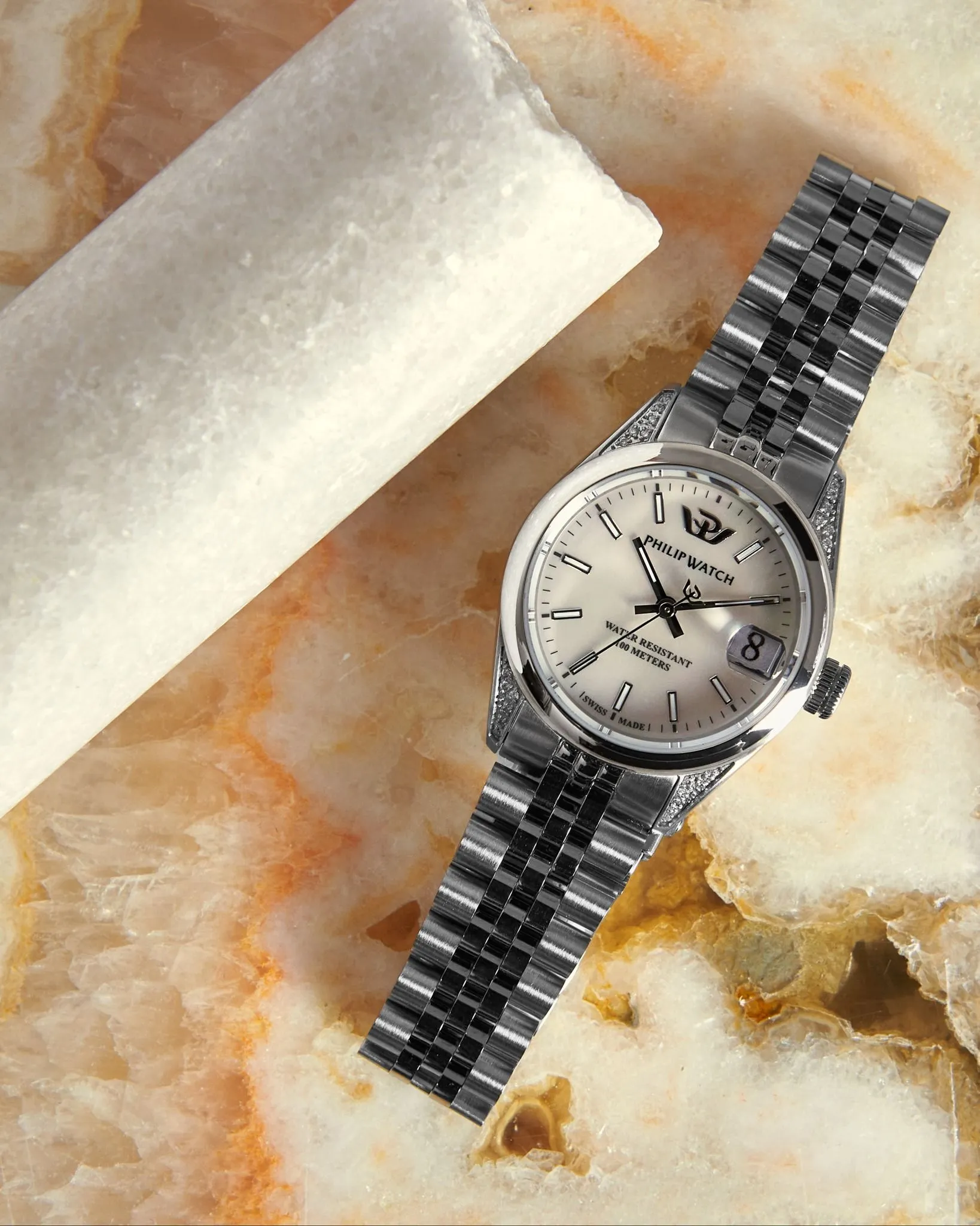 Orologio da polso in argento su una superficie decorata accanto a un oggetto cilindrico bianco.